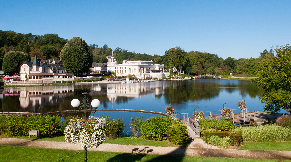 A white luxury hotel beside an attractive lake set amid woodland, with a clear blue sky